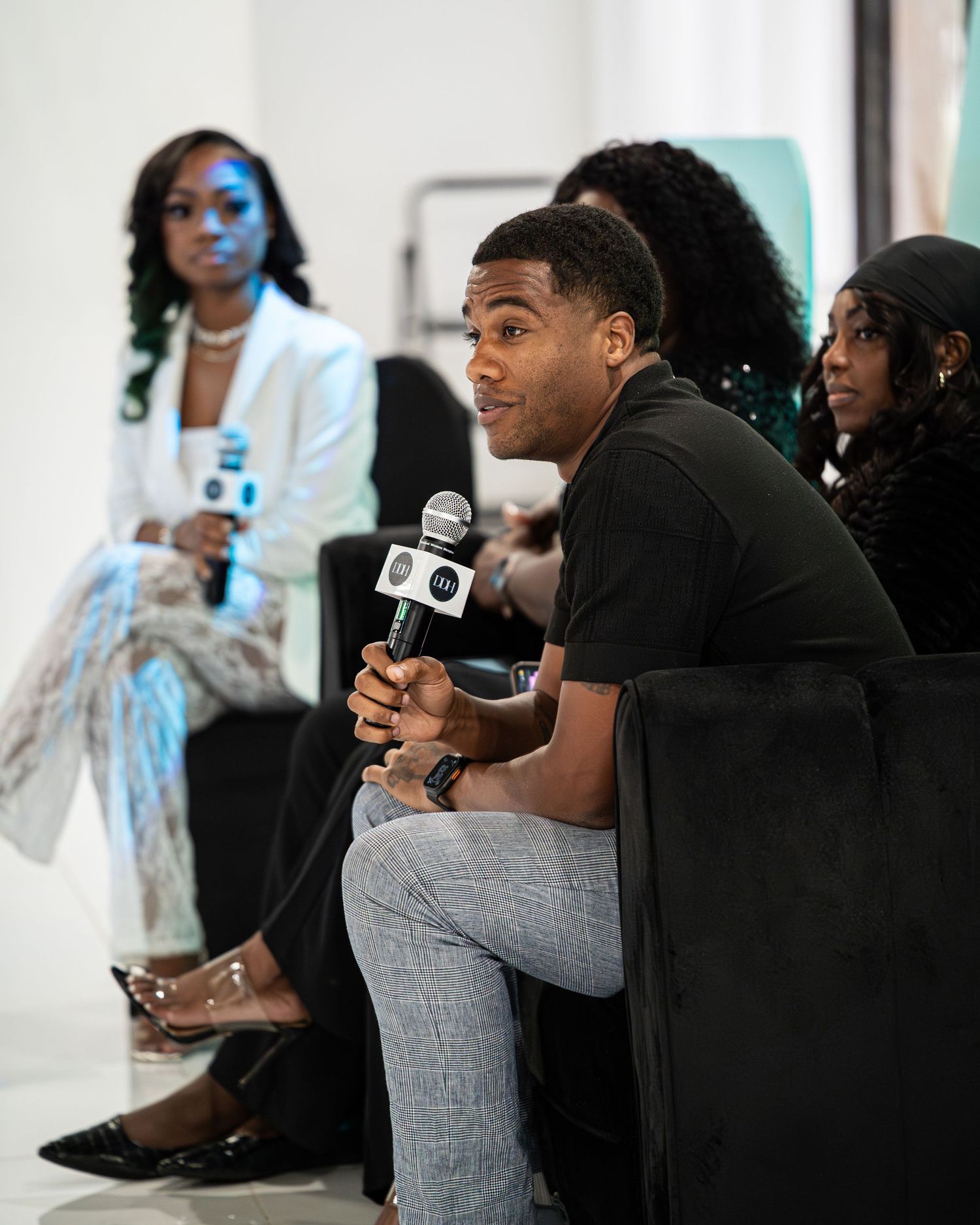 Young man speaks into microphone at panel event seated before diverse audience.