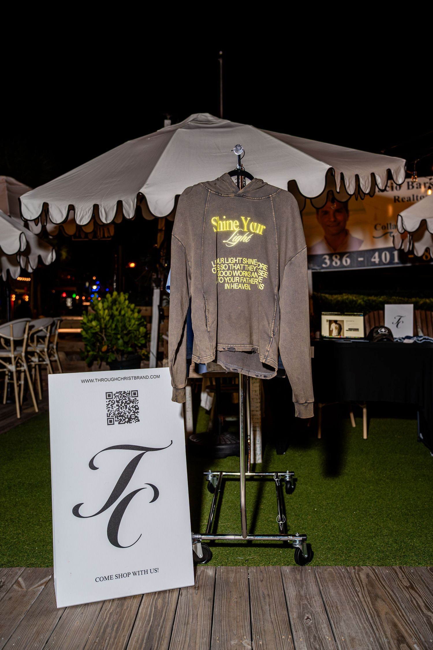 Brown Shine Your Light graphic sweatshirt on display rack with TC brand sign at night market.