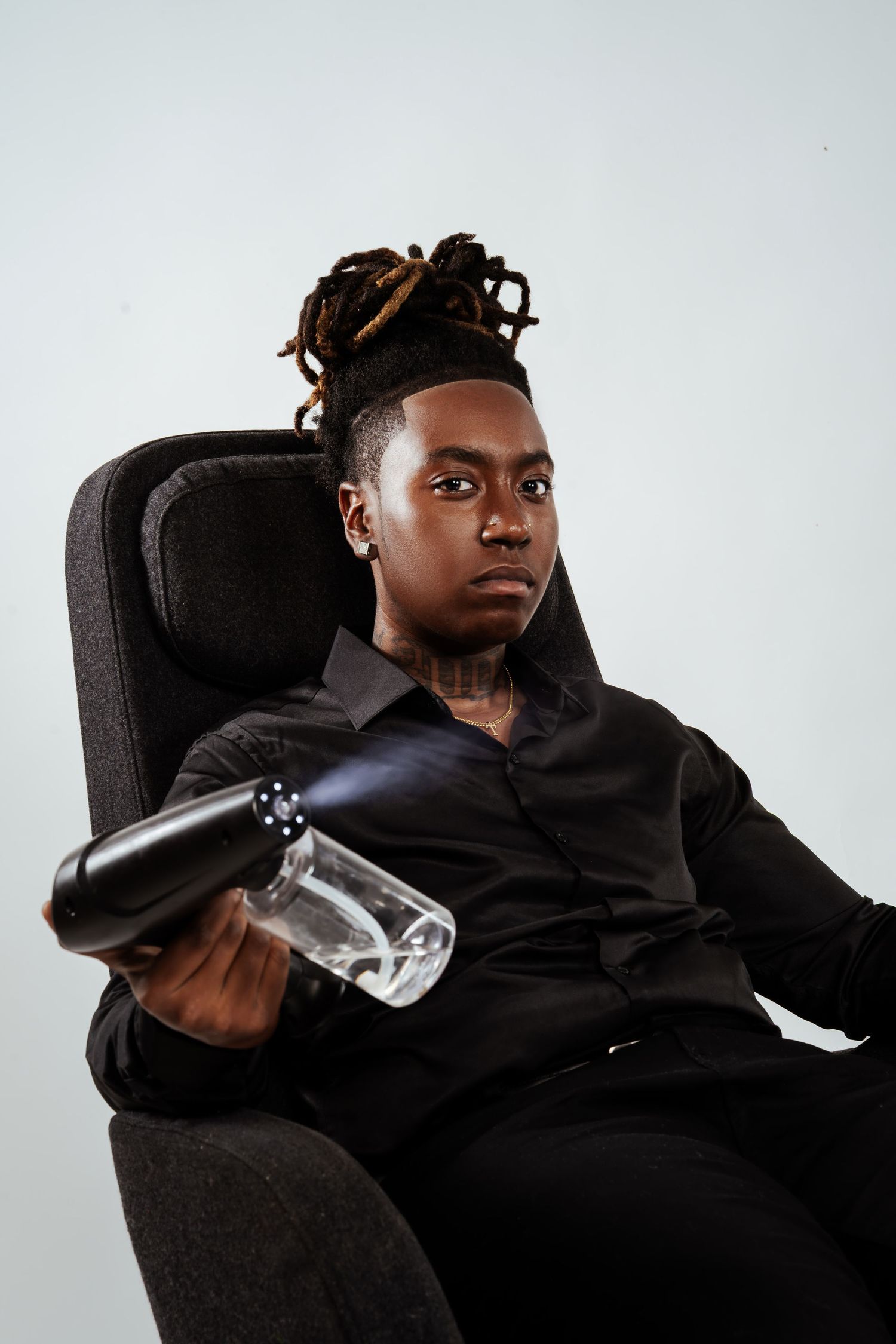 Young Black man with dreadlocks in black shirt seated in chair holding a glass bong with serious expression.