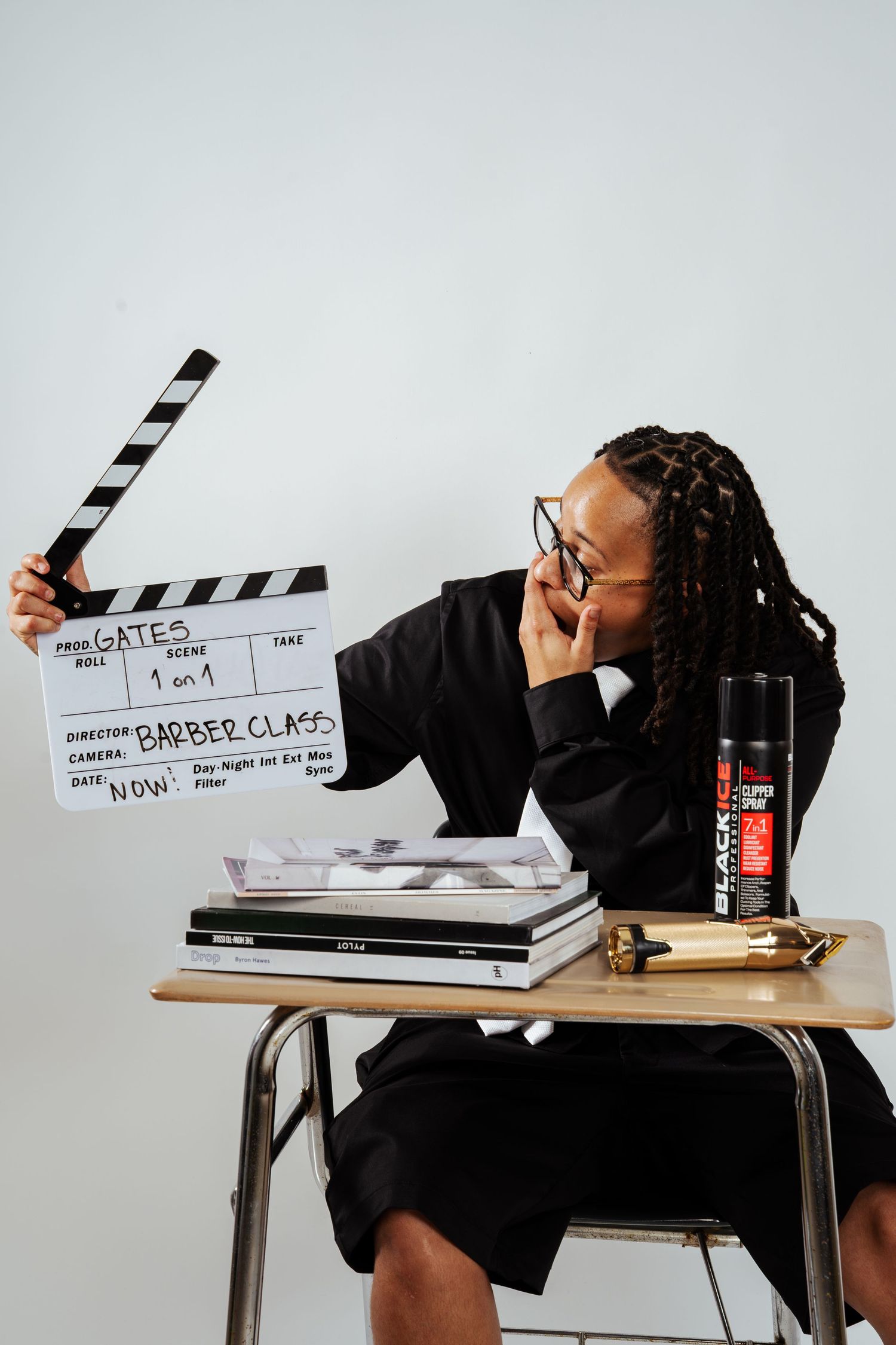 Person in black suit laughing while holding film clapperboard at school desk with books and Black Ice spray can.