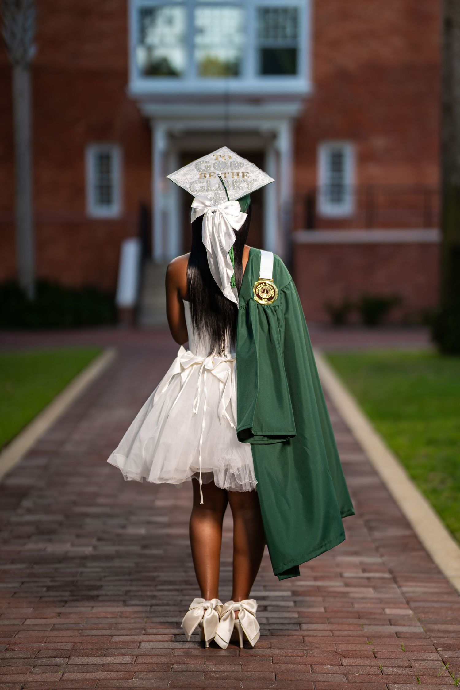 Graduate in white dress holding green gown stands on brick campus pathway facing historic building.