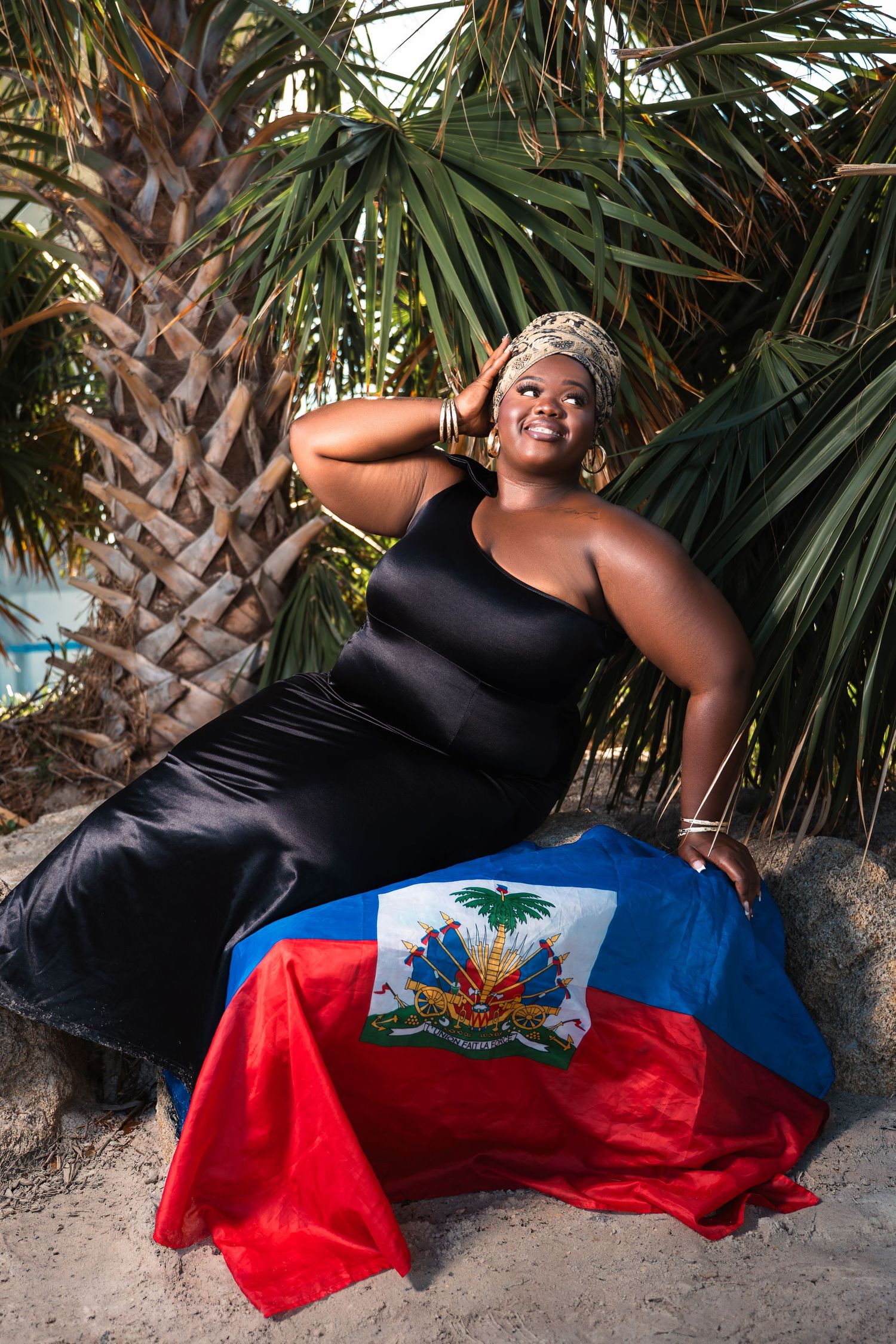 Smiling woman in black dress holding Haitian flag while sitting against palm trees on sandy beach.