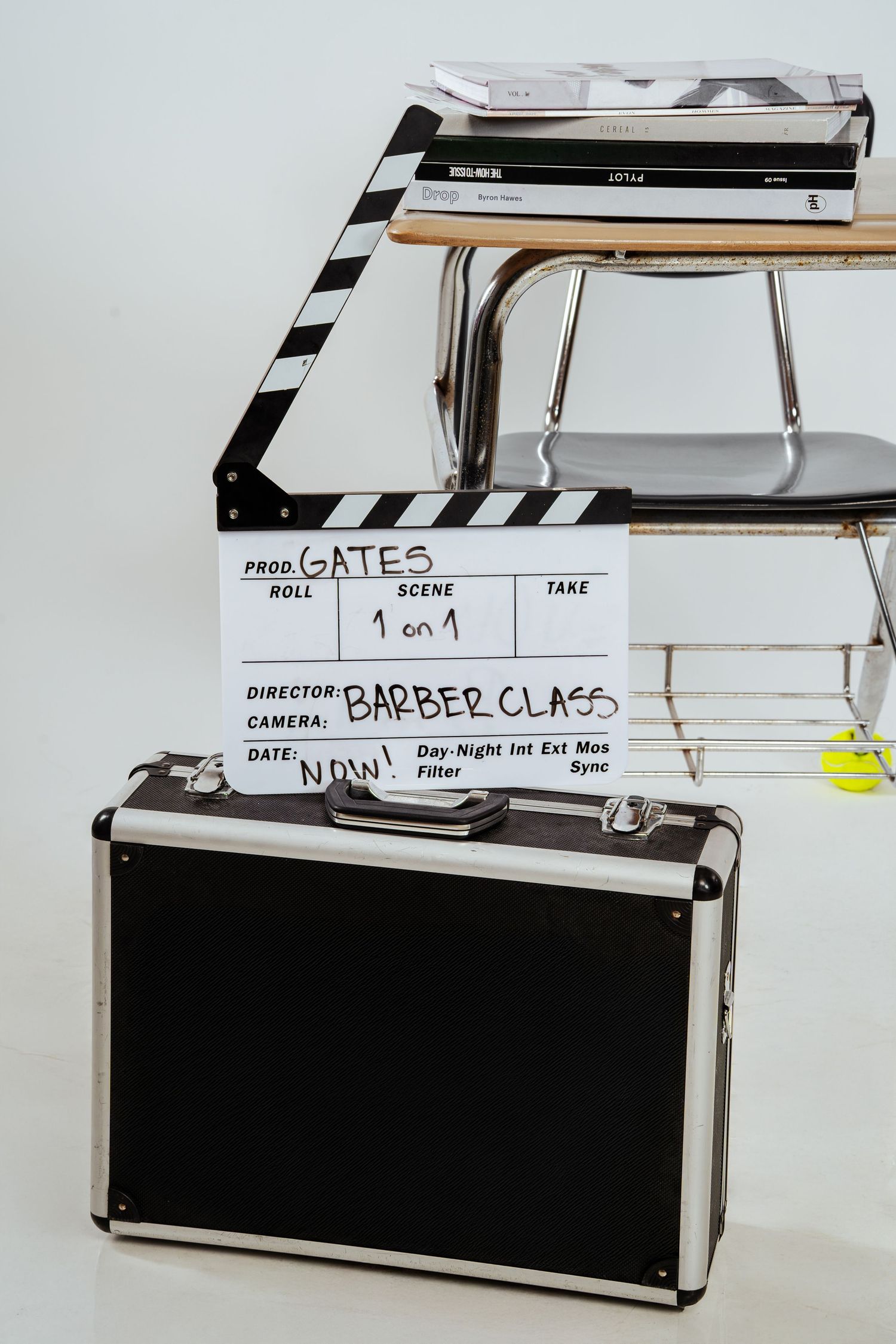 Film clapperboard labeled Gates Barber Class leaning against aluminum briefcase near school desk with books.