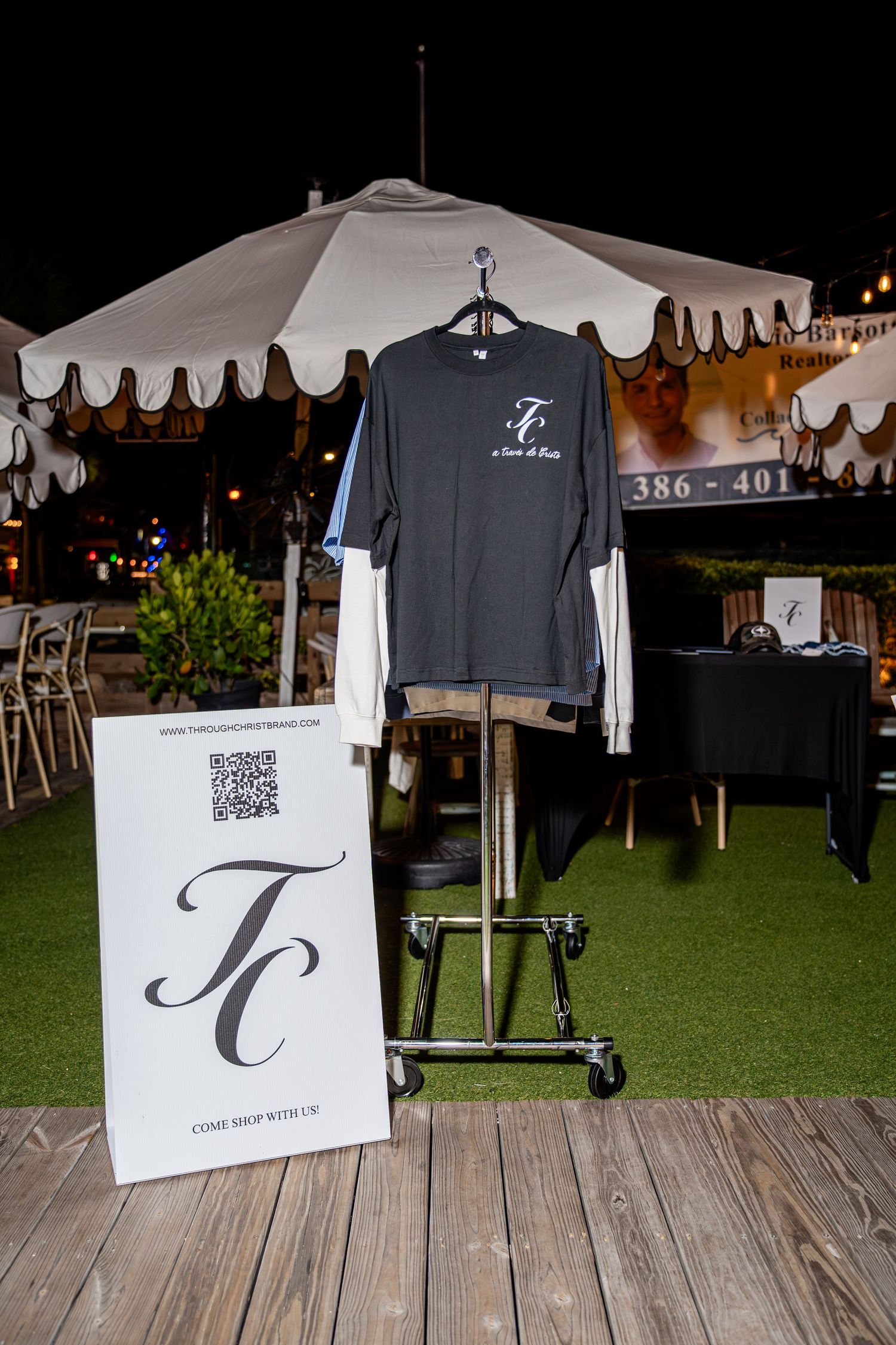 Dark gray TC a favor de Cristo t-shirt layered on rack with TC brand sign at outdoor event.