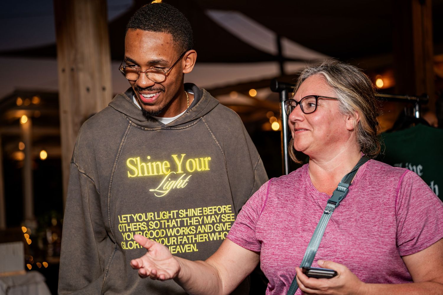 Man in Shine Your Light hoodie laughing with a woman at an outdoor nighttime event.