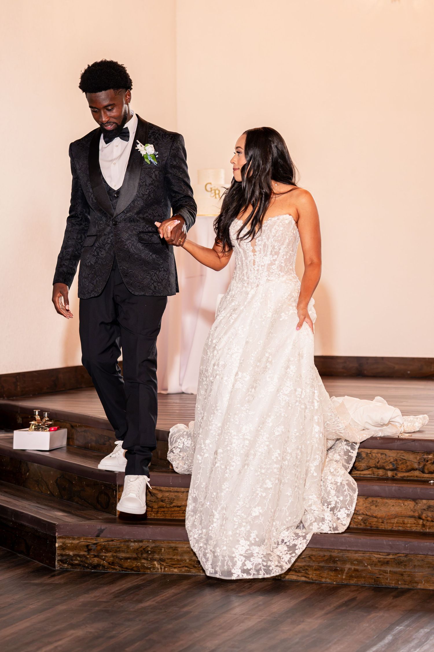 Bride and groom descend wooden steps hand in hand, groom in black tuxedo, bride in white lace wedding gown.