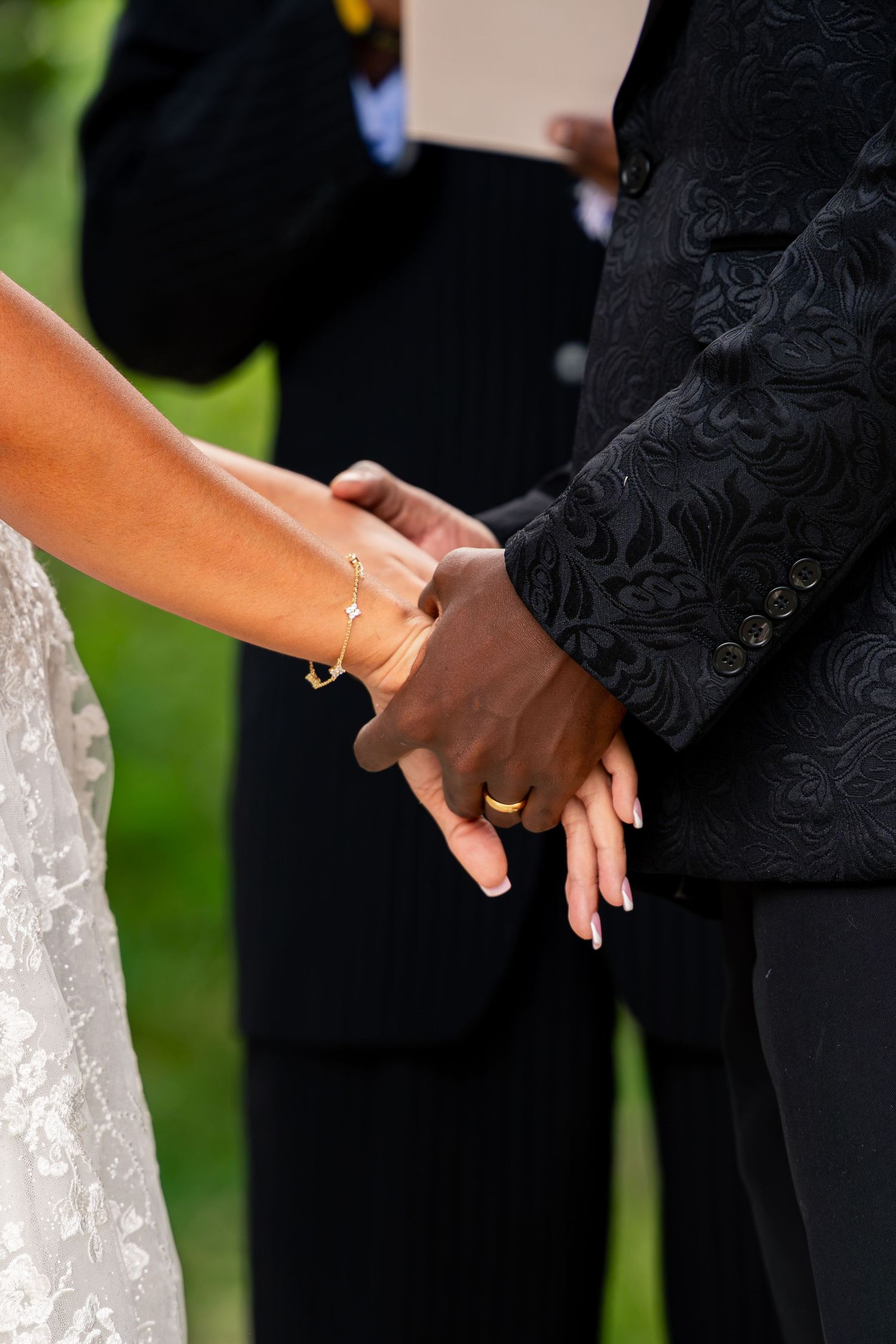 Bride and groom holding hands during outdoor wedding ceremony, bride wearing gold bracelet.