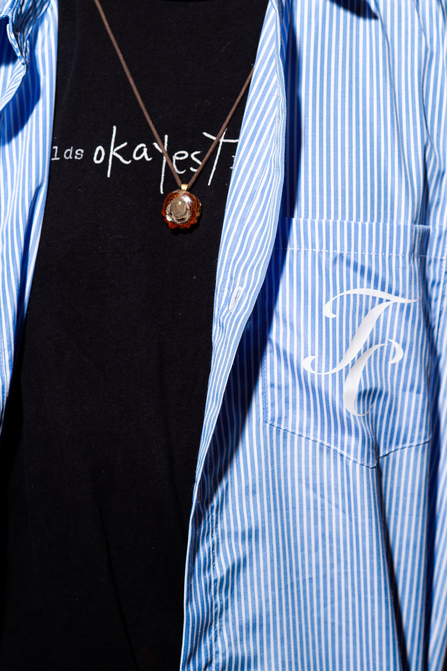 Close-up of black graphic tee with necklace under blue striped shirt with monogram pocket.