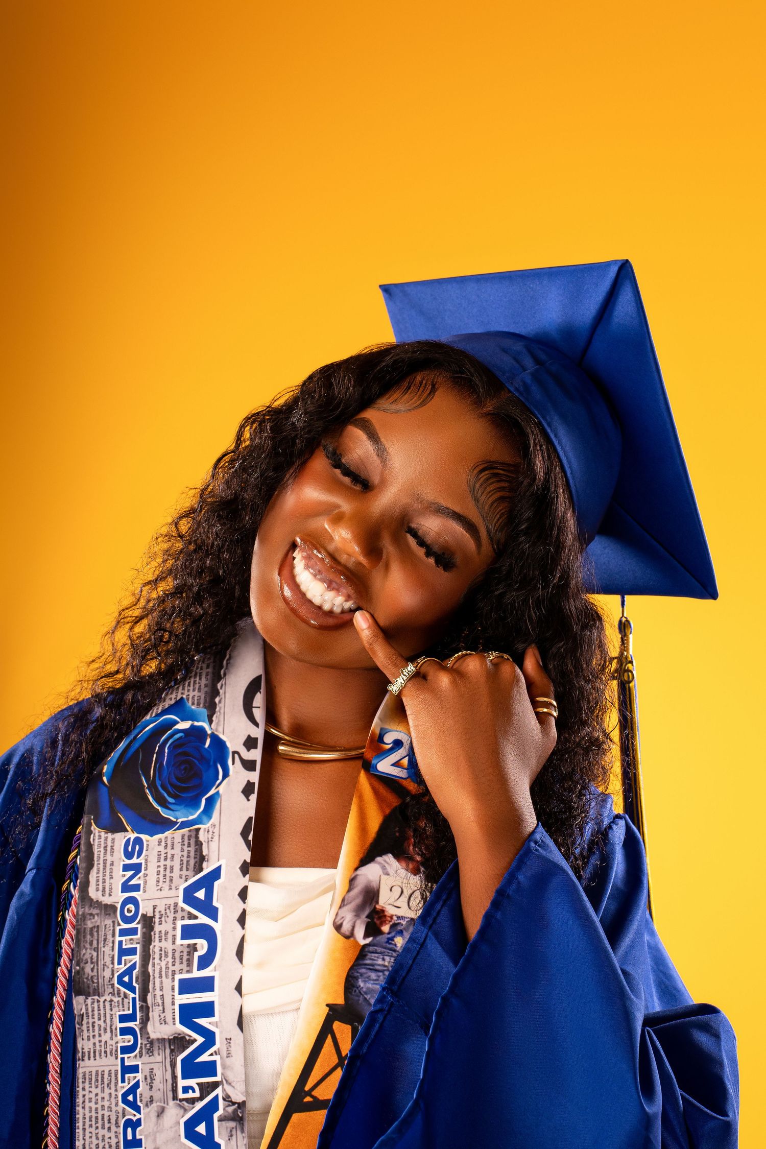 Happy graduate in blue cap and gown smiling warmly against bright yellow studio background.