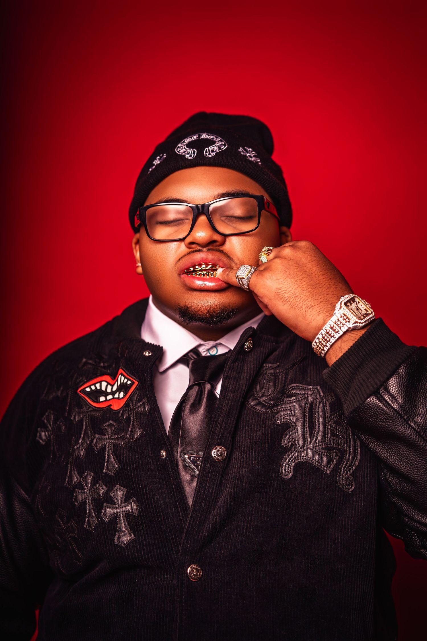 Man in black jacket with cross patches and gold grillz posing against vibrant red background.