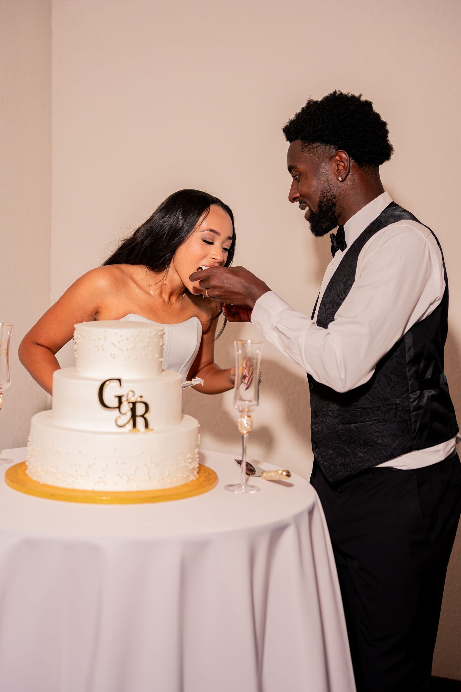 Groom feeds bride a bite of white three-tier wedding cake with G&R monogram topper.