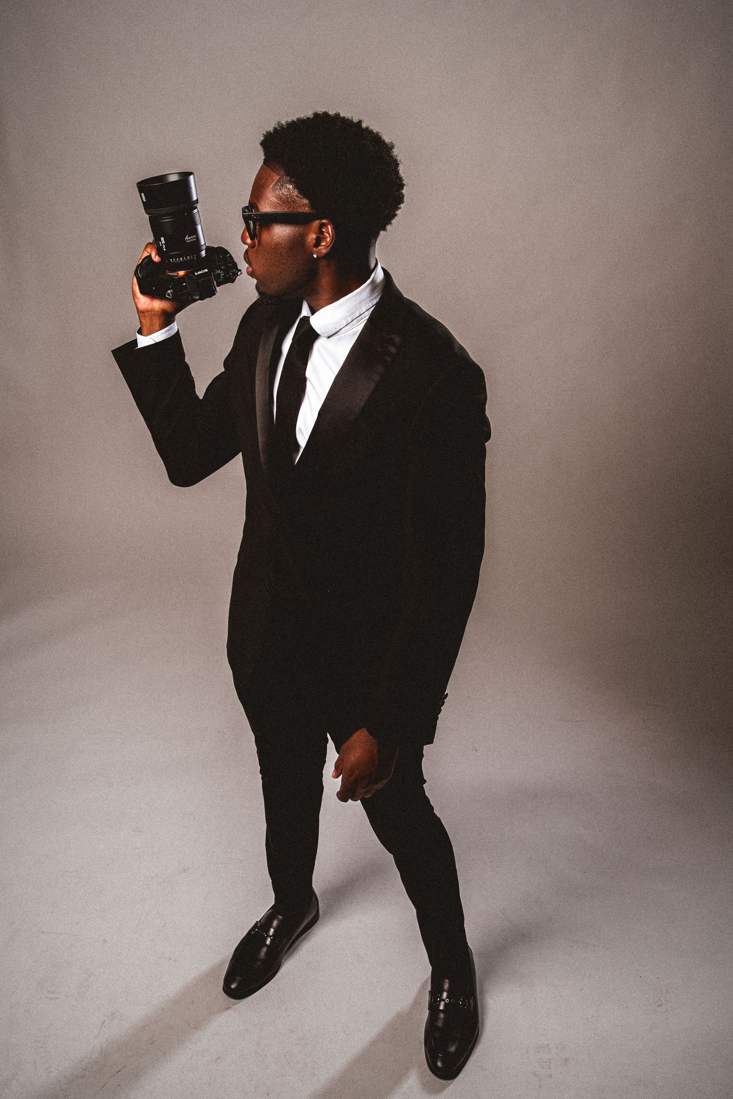 Young Black man in black tuxedo suit photographing with a professional camera on gray background.