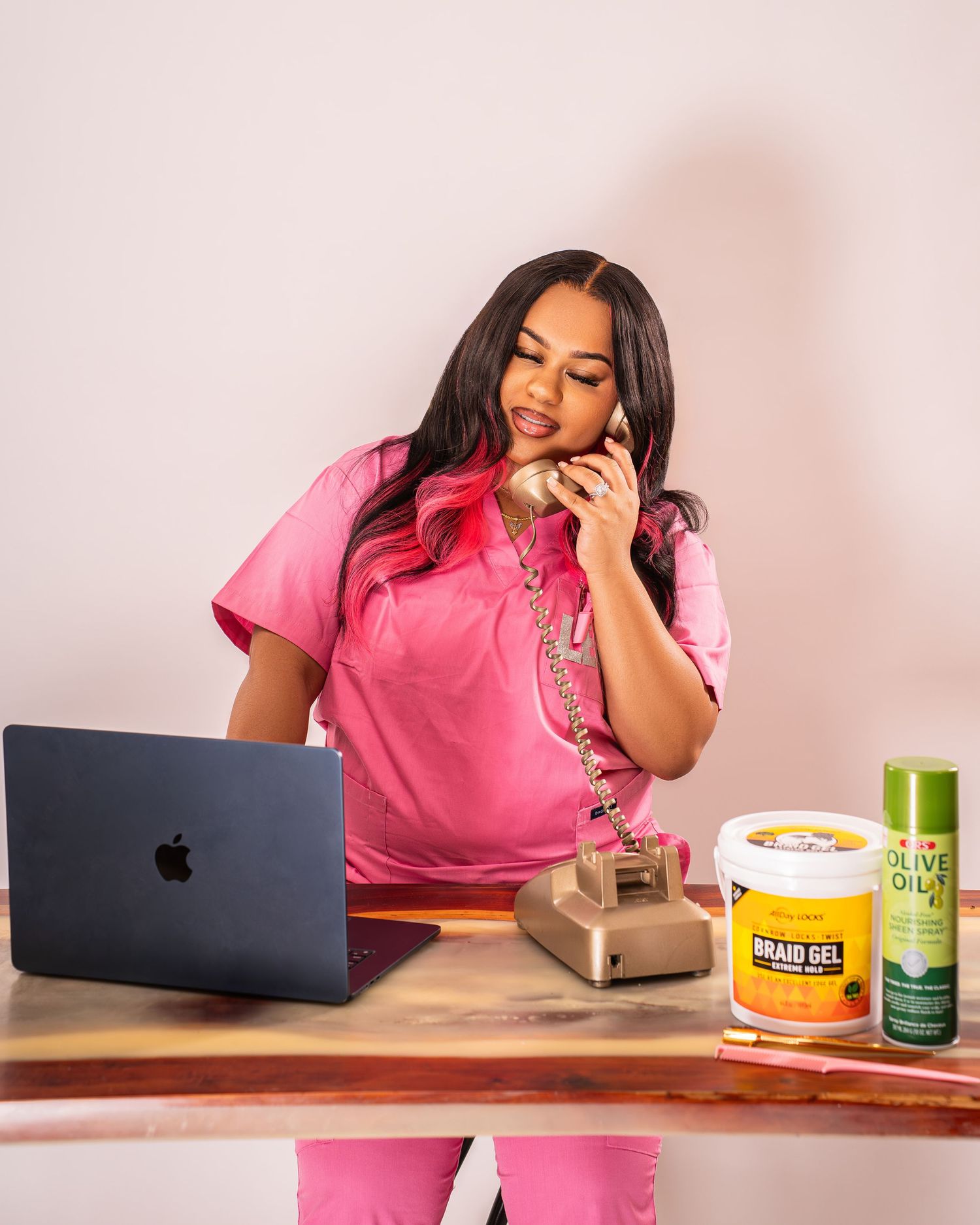 Woman in pink outfit talking on vintage phone at desk with MacBook and hair care products.