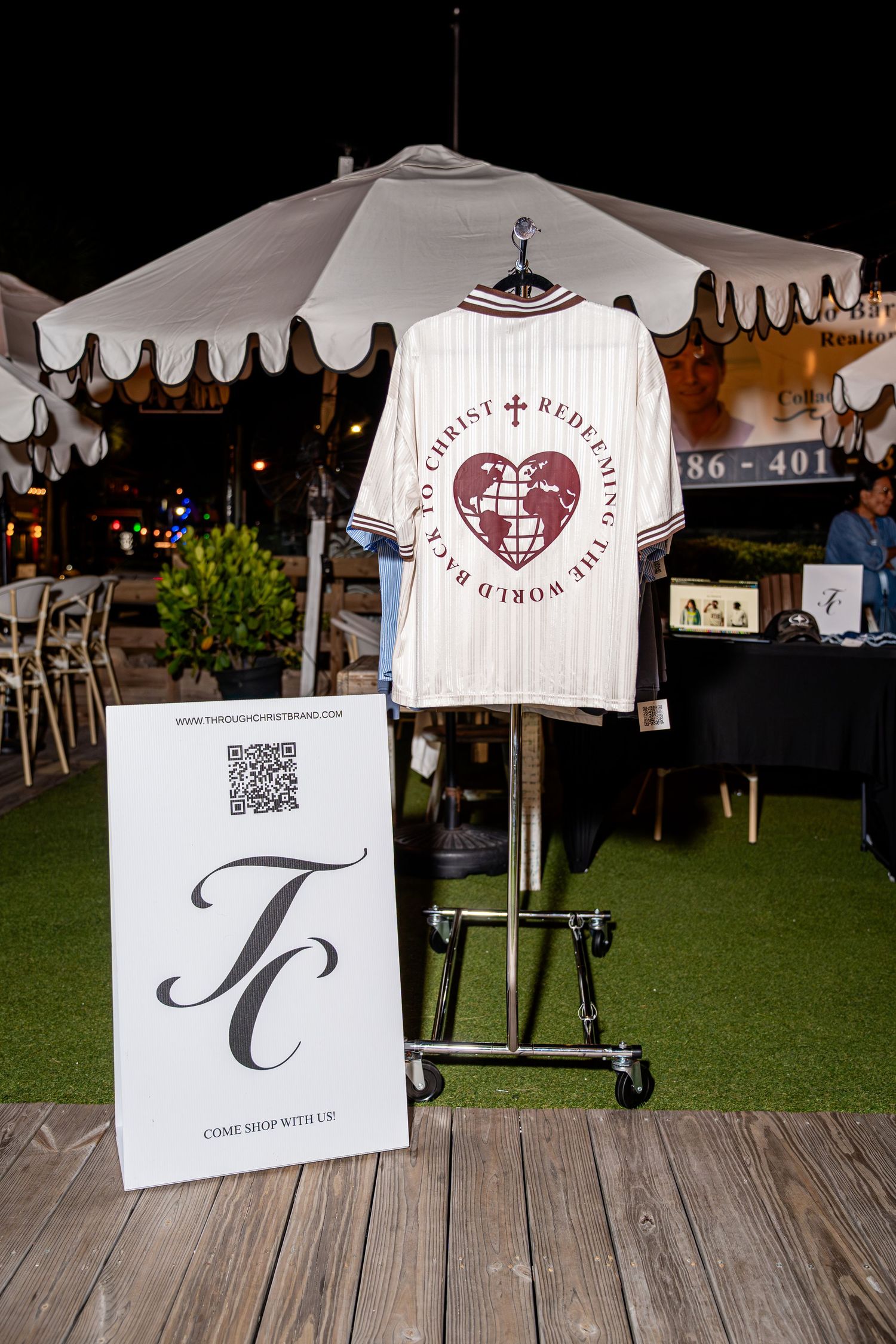 White pinstripe Through Christ Redeeming the World baseball jersey on outdoor display rack at night.