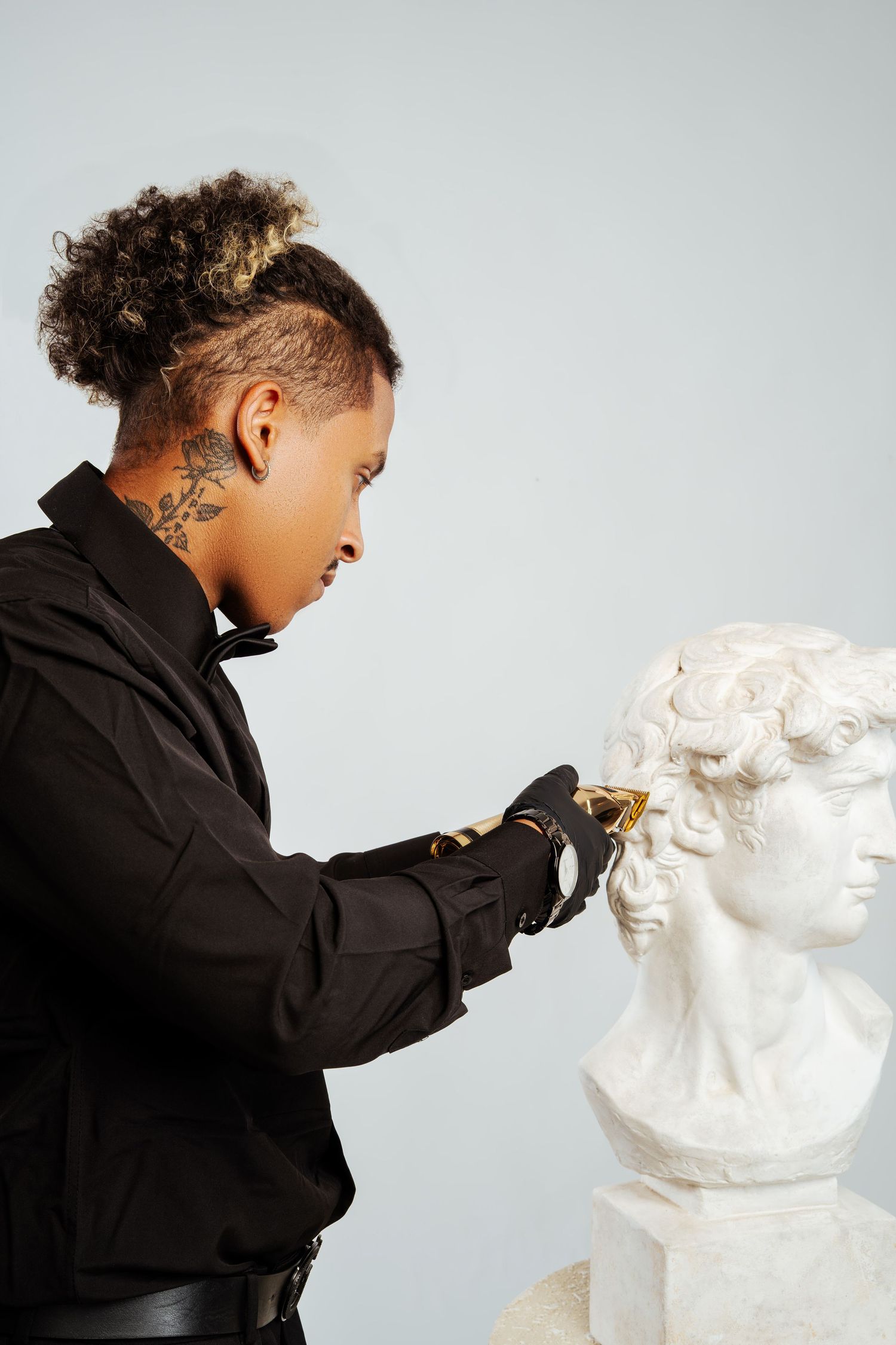 Barber with neck tattoos using gold clippers on a classical white marble bust sculpture.