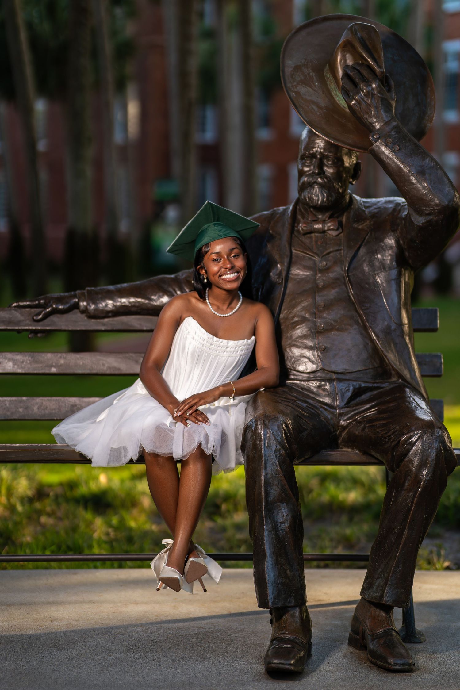 Smiling graduate in white dress and green cap poses beside bronze statue on campus bench outdoors.