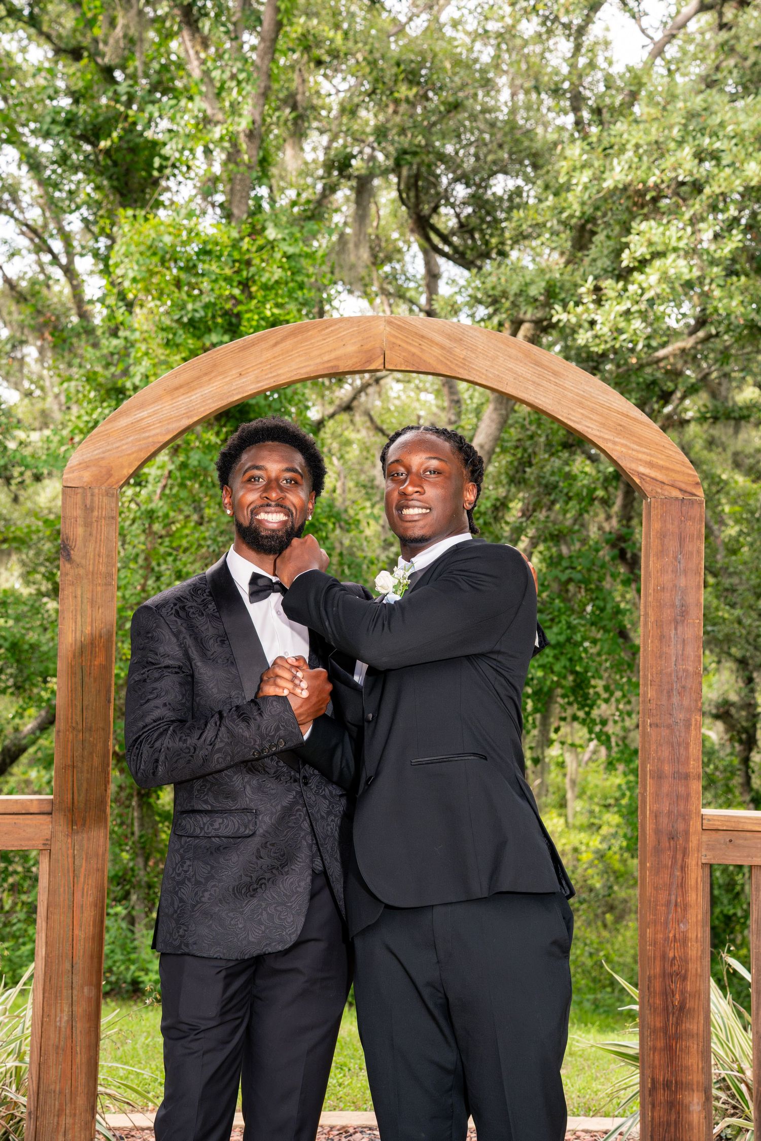 Two men in black tuxedos laughing together under a wooden arch at an outdoor wedding ceremony.