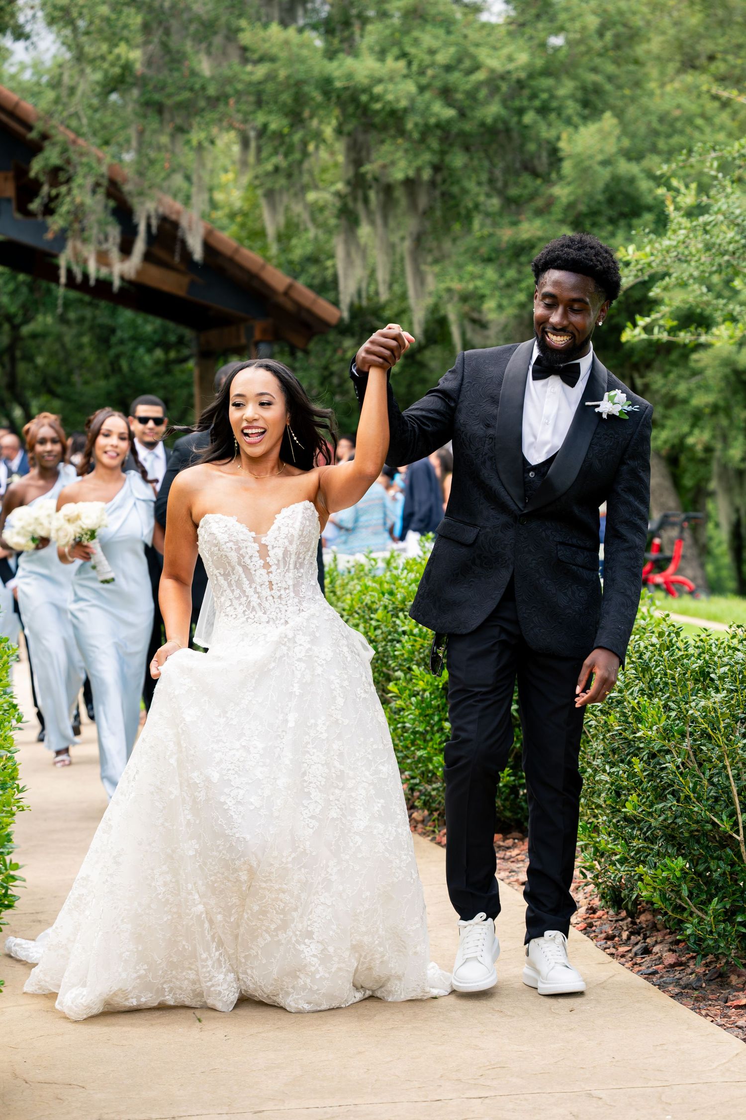 Joyful newlywed couple walks hand-in-hand down outdoor wedding path, bride in lace ballgown, groom in black tuxedo.