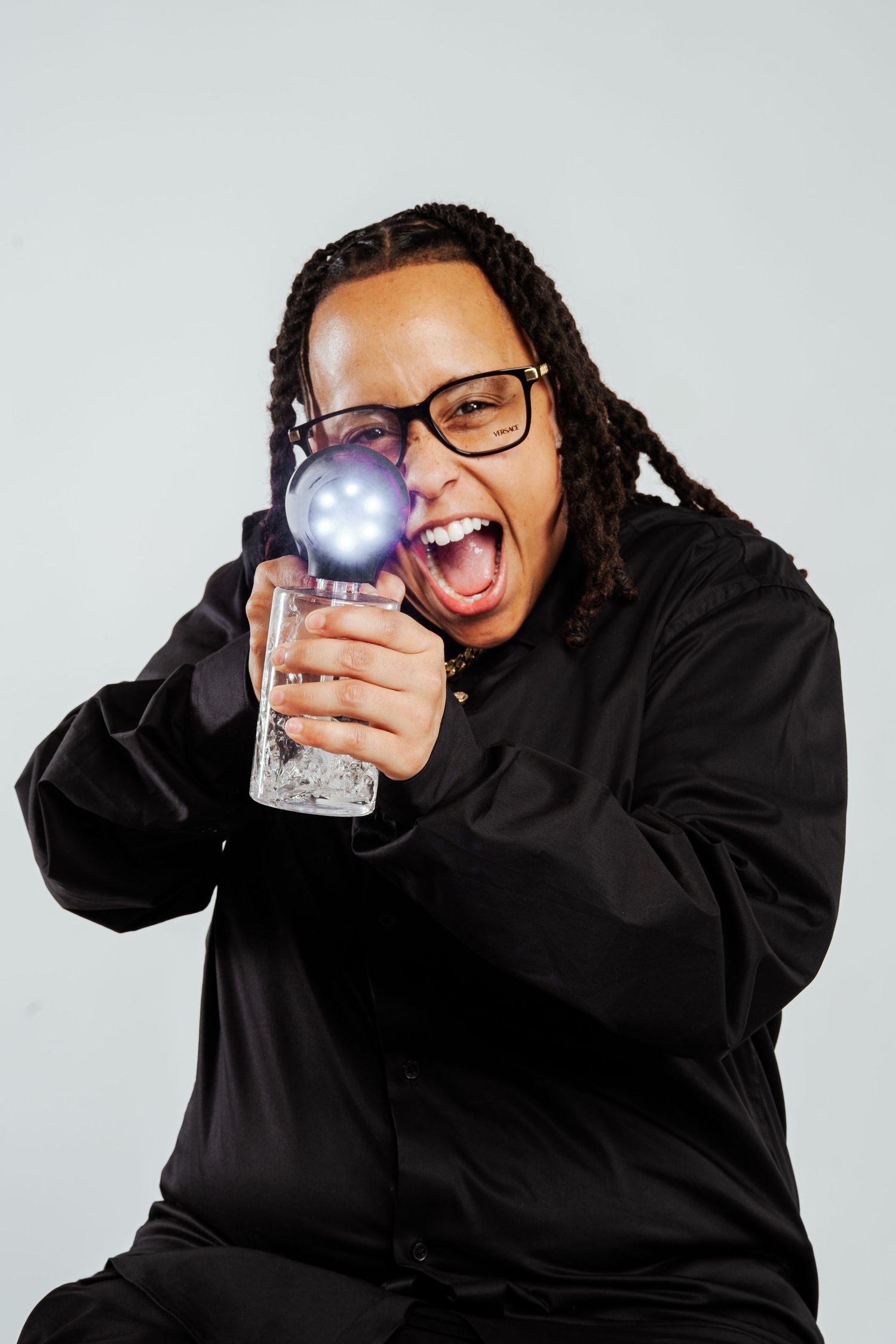 Person with glasses and dreadlocks pointing glowing glass cup toward camera with open mouth.