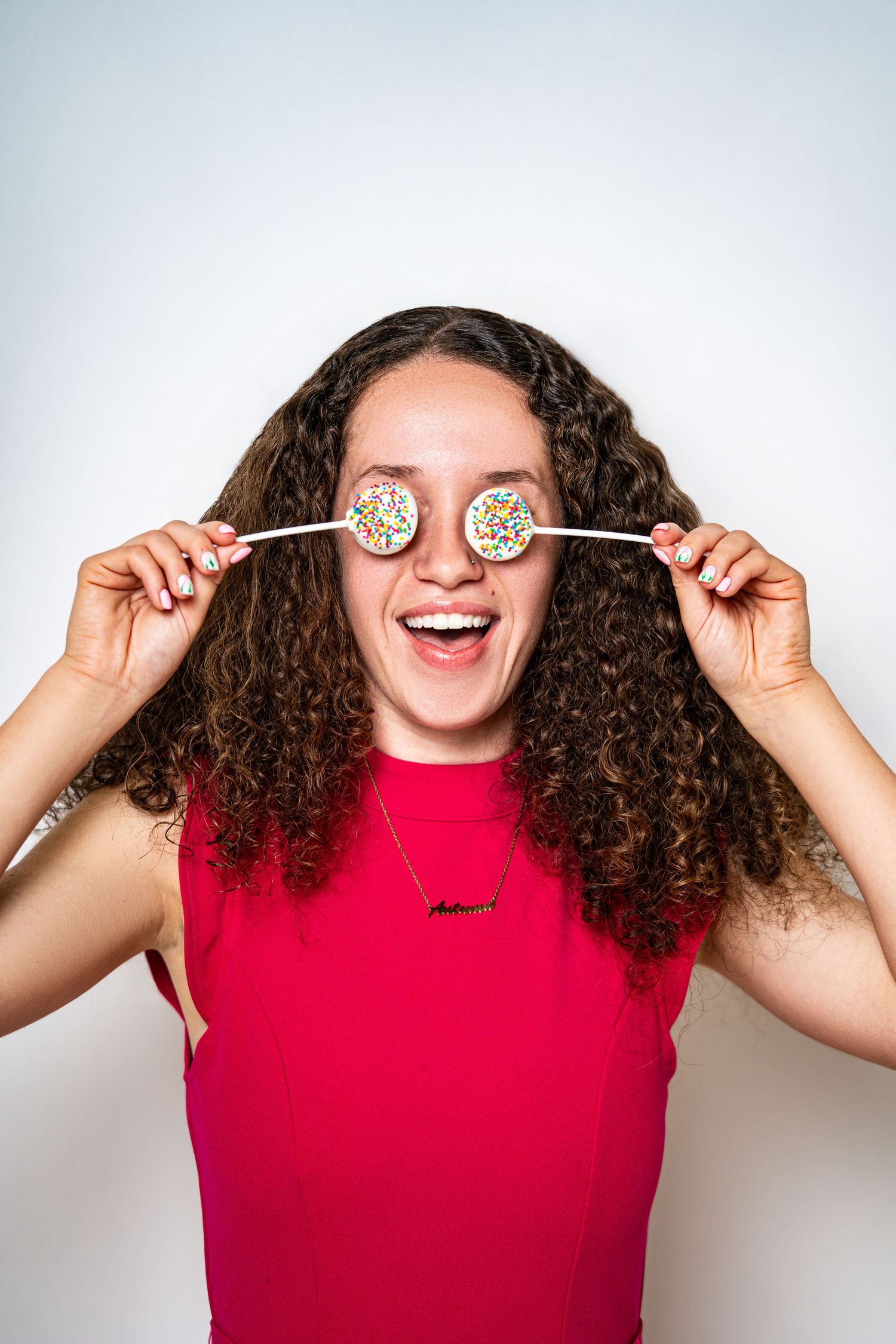 Joyful young woman with curly hair covering eyes with two colorful sprinkle cake pops in red dress.