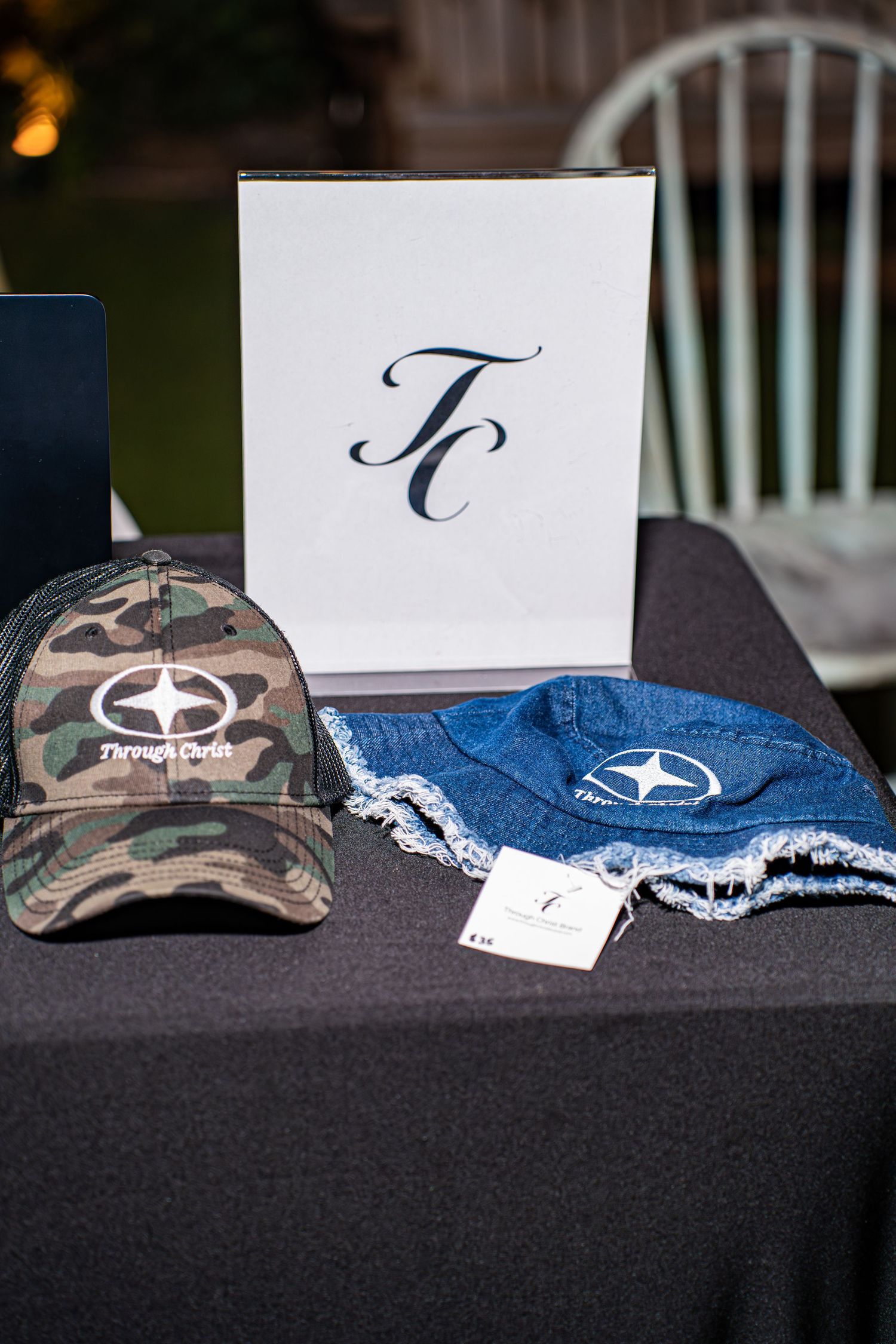 Camo and denim hats with TC logo displayed on black table at Christian merchandise booth.