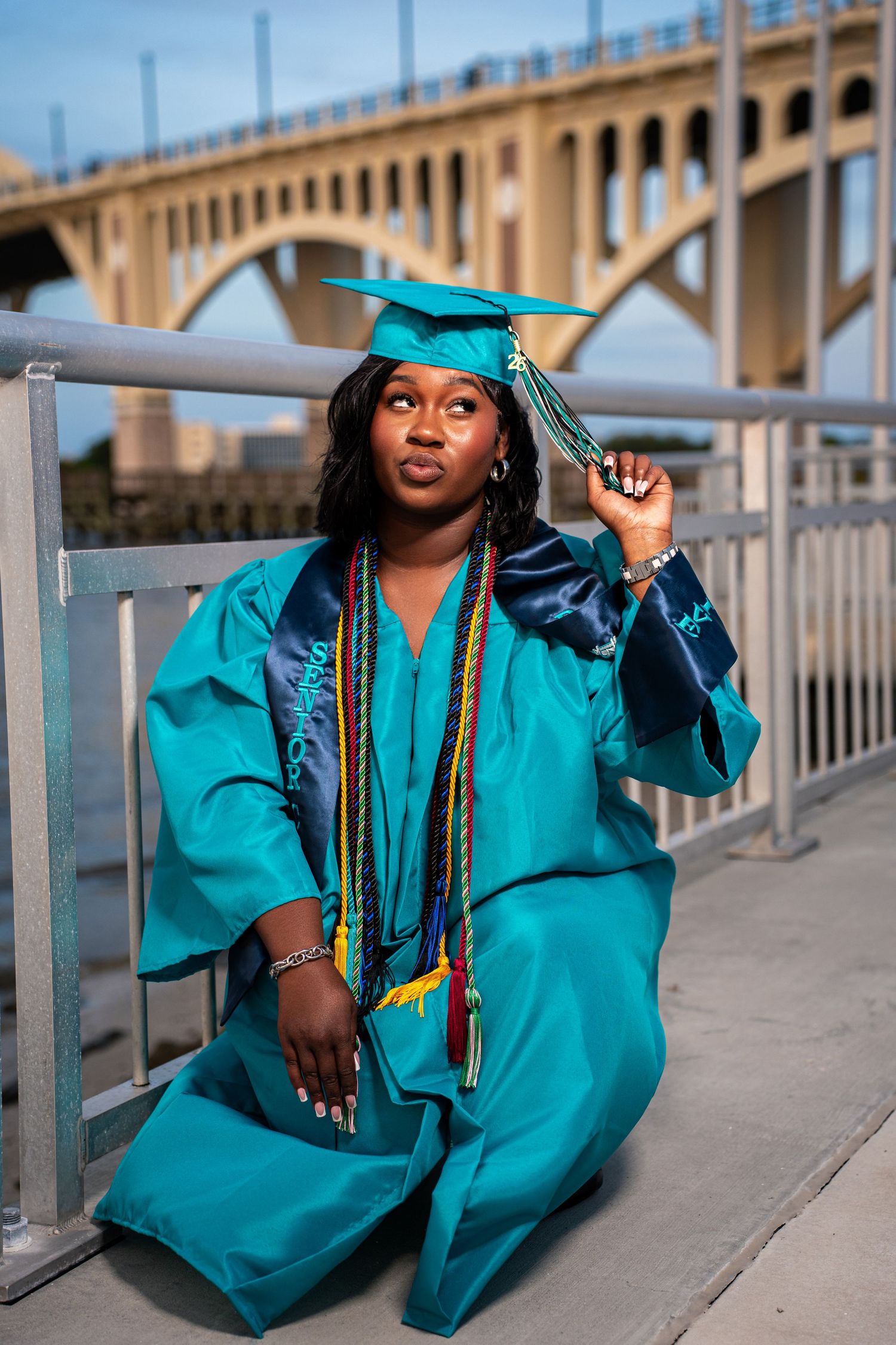 Graduate in teal cap and gown sits by bridge railing holding tassel, looking thoughtfully upward.