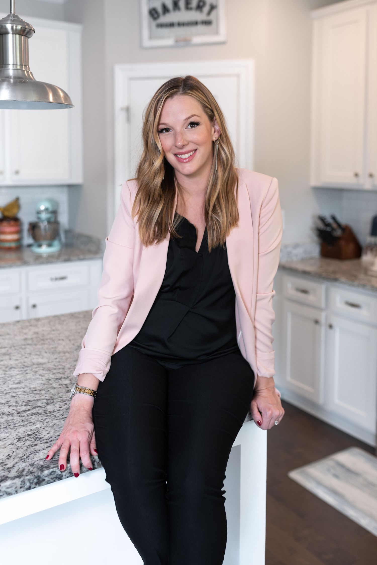 Woman in pink blazer and black outfit sitting on kitchen counter with modern white cabinets.