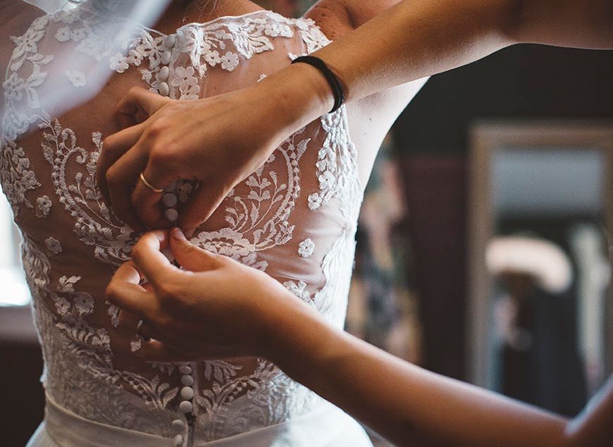Photograph of a bridesmaid buttoning up the back of the bride's dress.