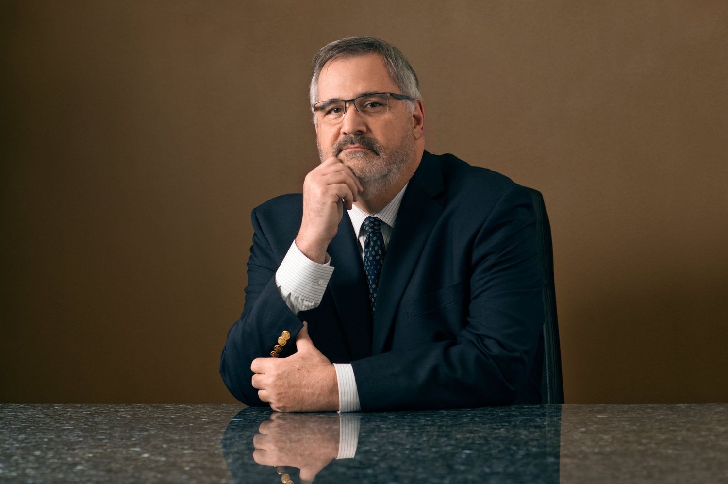 Professional businessman in dark suit sitting at marble table with hand on chin in thoughtful pose.