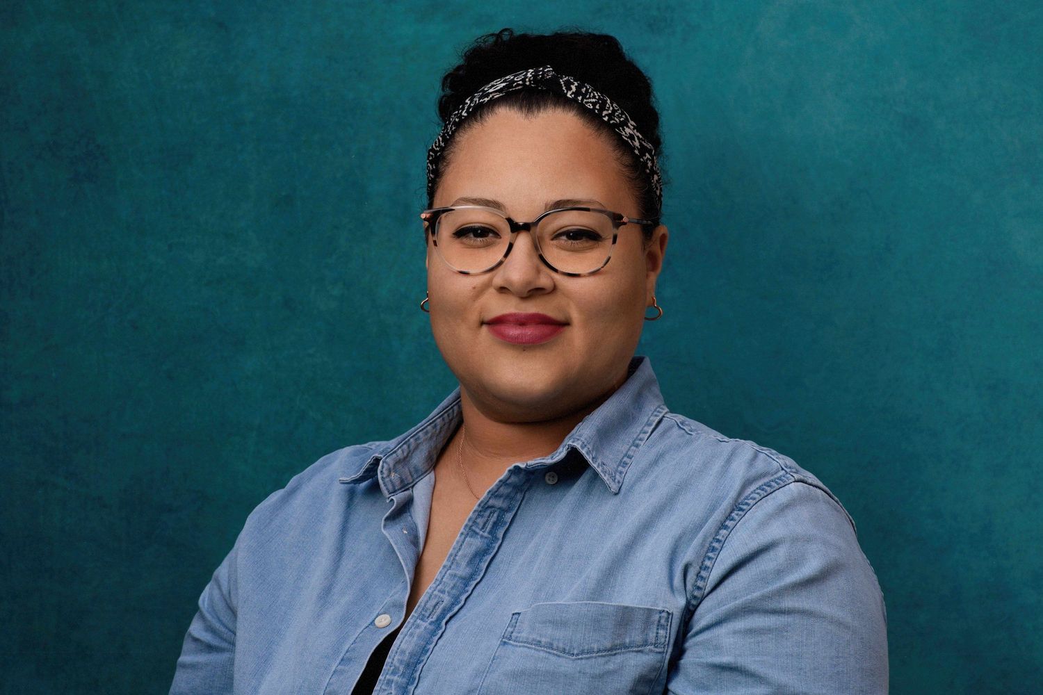 Woman with glasses and patterned headband poses in denim shirt against teal background.