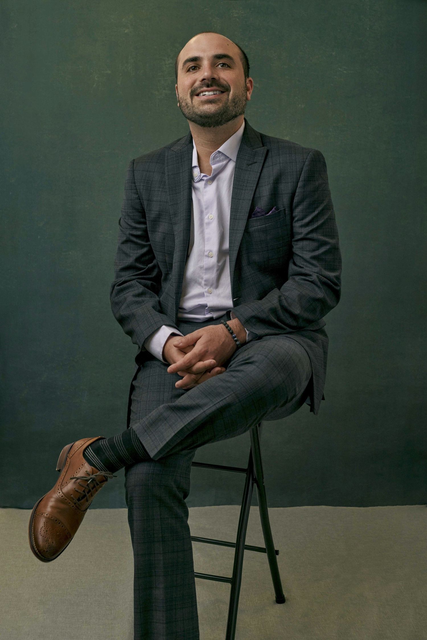 Smiling young man in grey plaid suit seated on stool with legs crossed against dark green background.