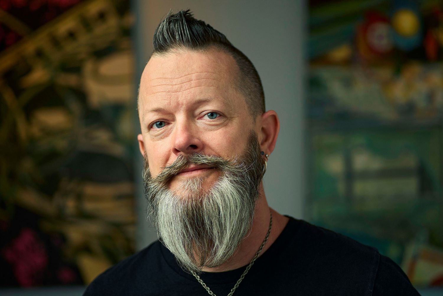 Man with stylish salt-and-pepper beard and mohawk hairstyle poses in front of colorful artwork background.