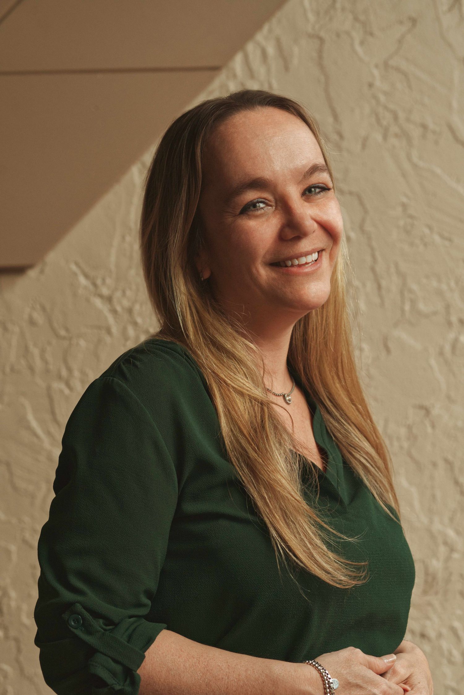 Smiling woman with long blonde hair wearing green blouse posing against textured wall in warm lighting.
