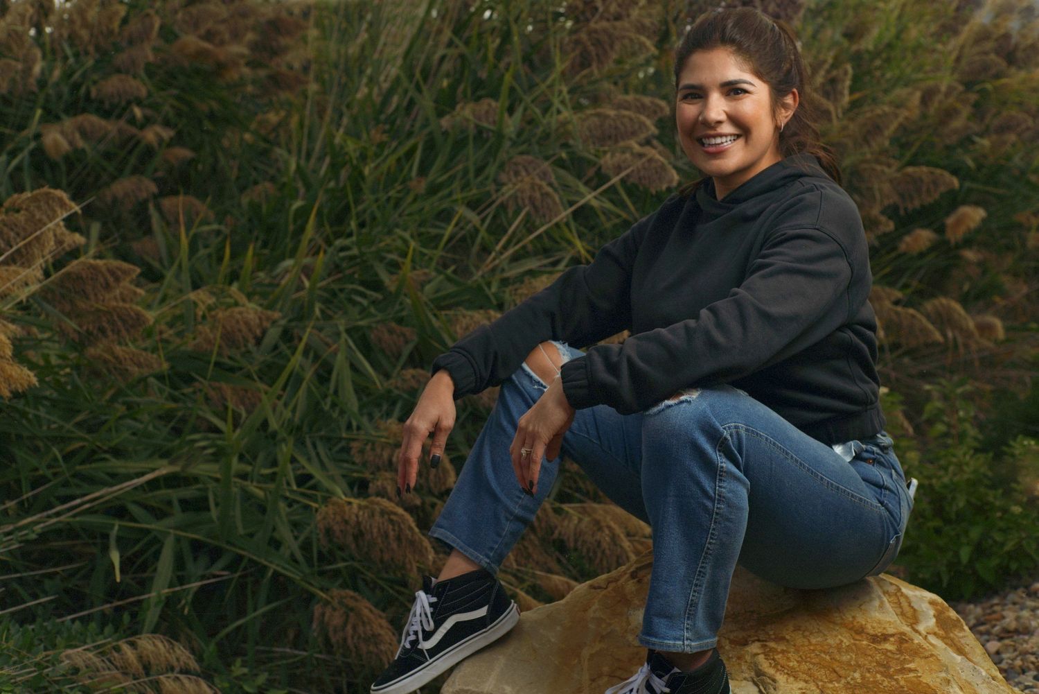 Smiling woman in black hoodie and jeans sits on rock outdoors in nature.