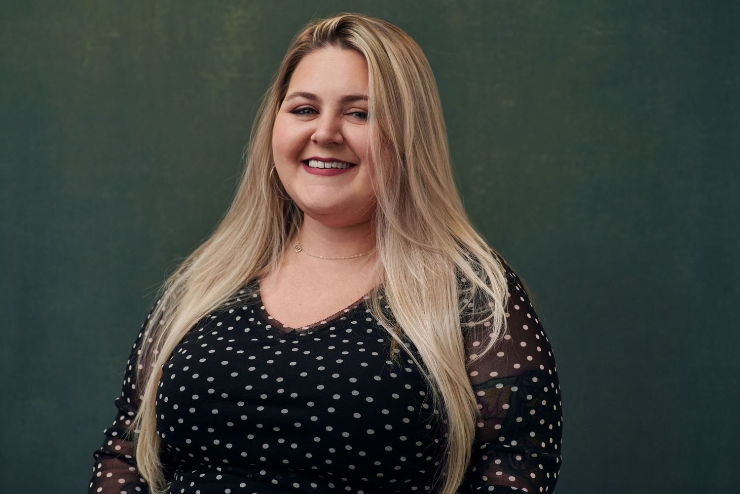 Smiling blonde woman in black polka dot dress poses against dark green background.