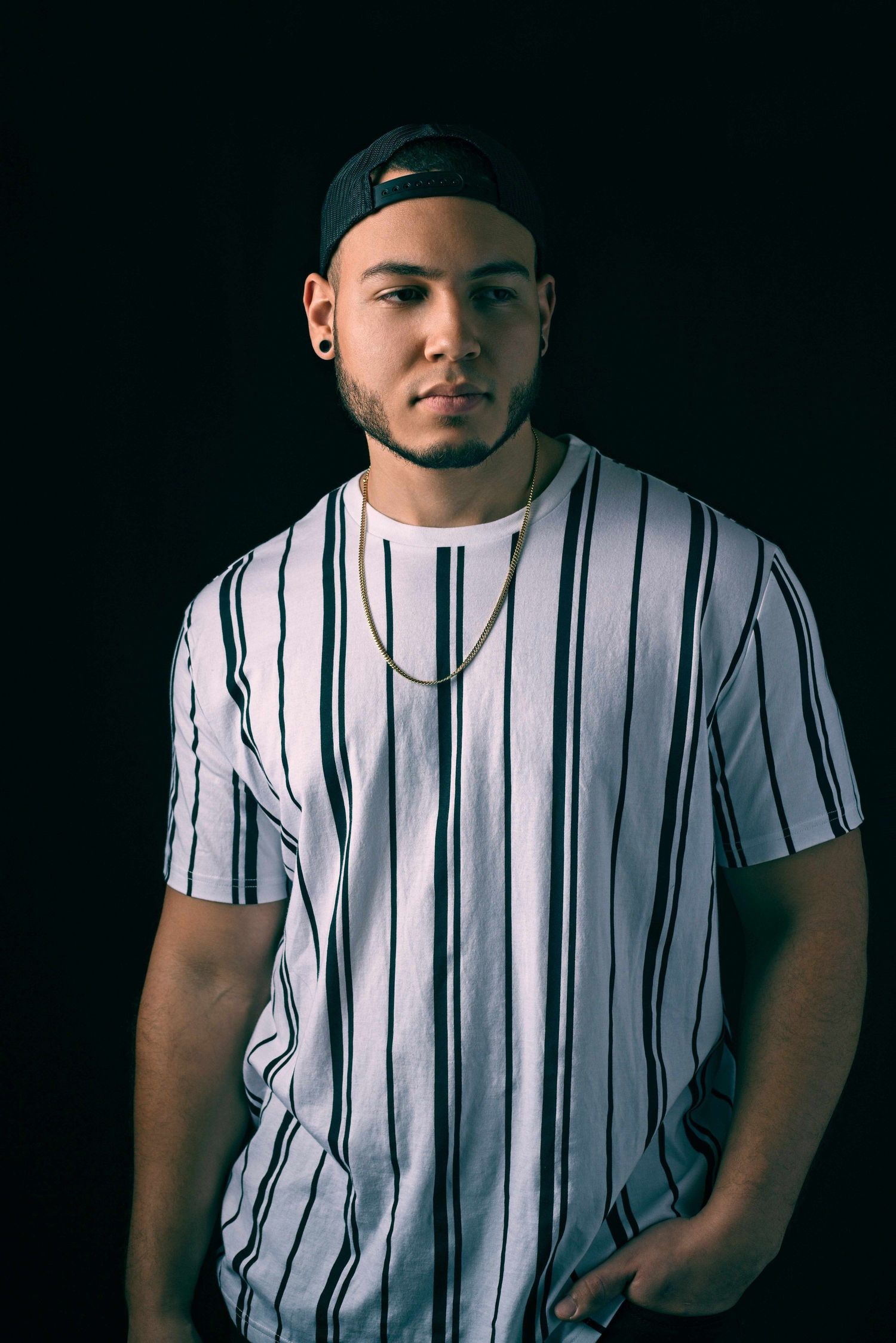 Young man in striped white tee and backwards cap poses against black background.