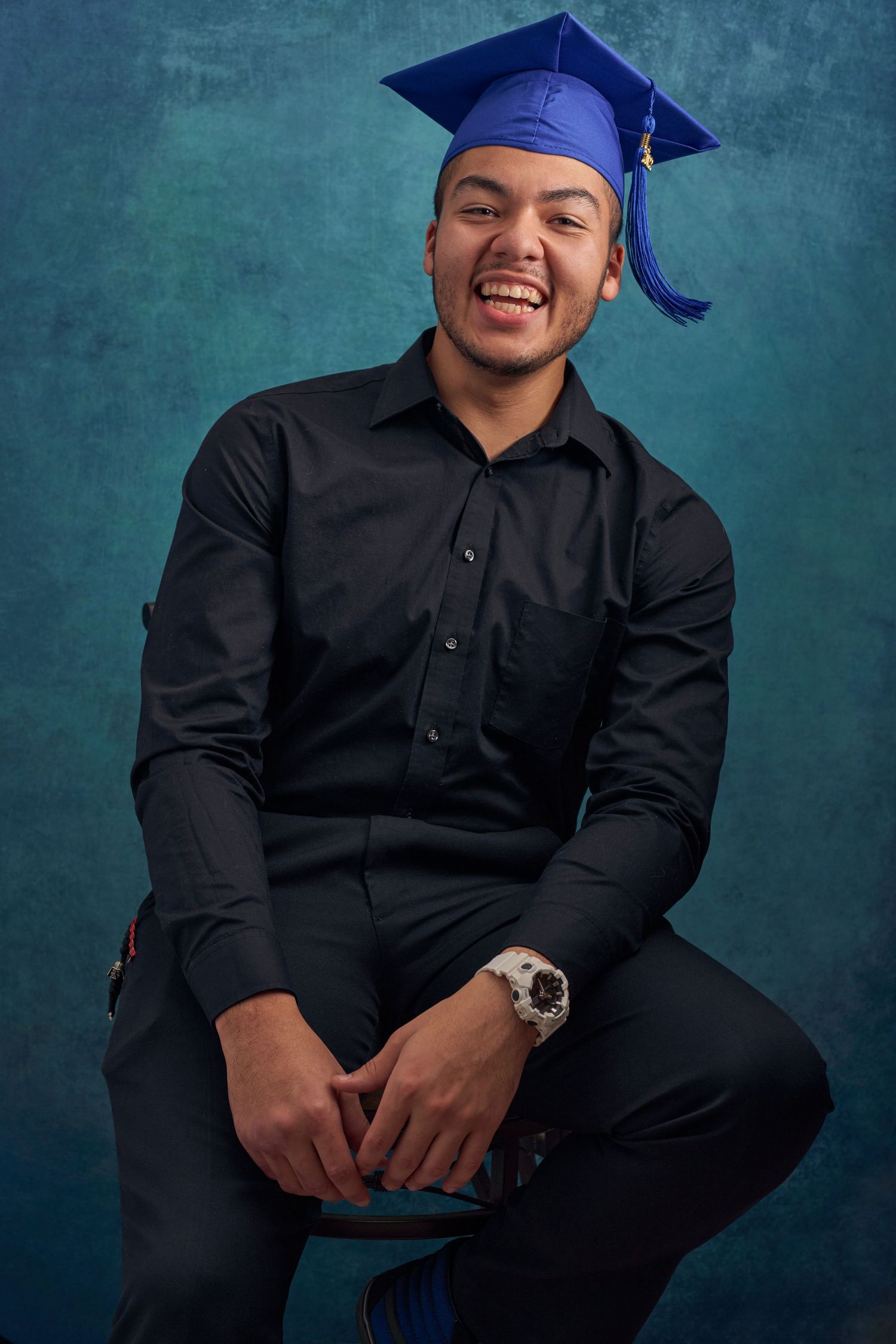 Smiling young man in black shirt and blue graduation cap sits proudly on stool against teal studio background.