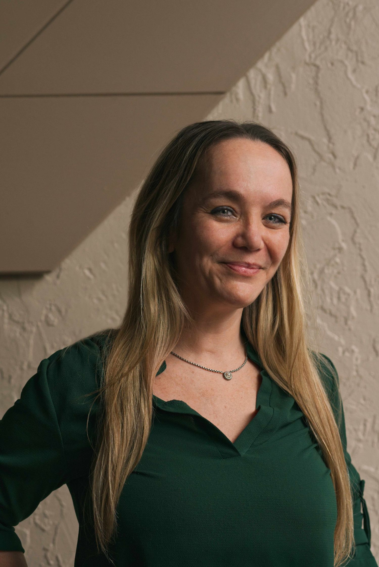 Woman with long blonde hair in green top smiles outdoors against textured wall.