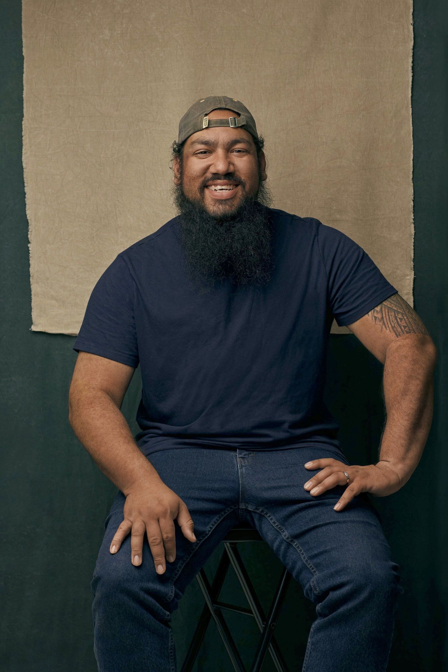 Bearded man in navy tee and backwards cap sits smiling on stool indoors.
