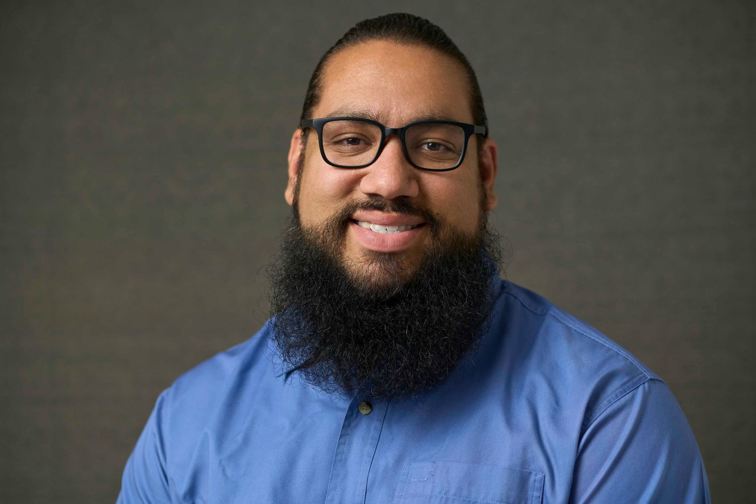 Bearded man with glasses smiles in blue button-up shirt against gray studio background.