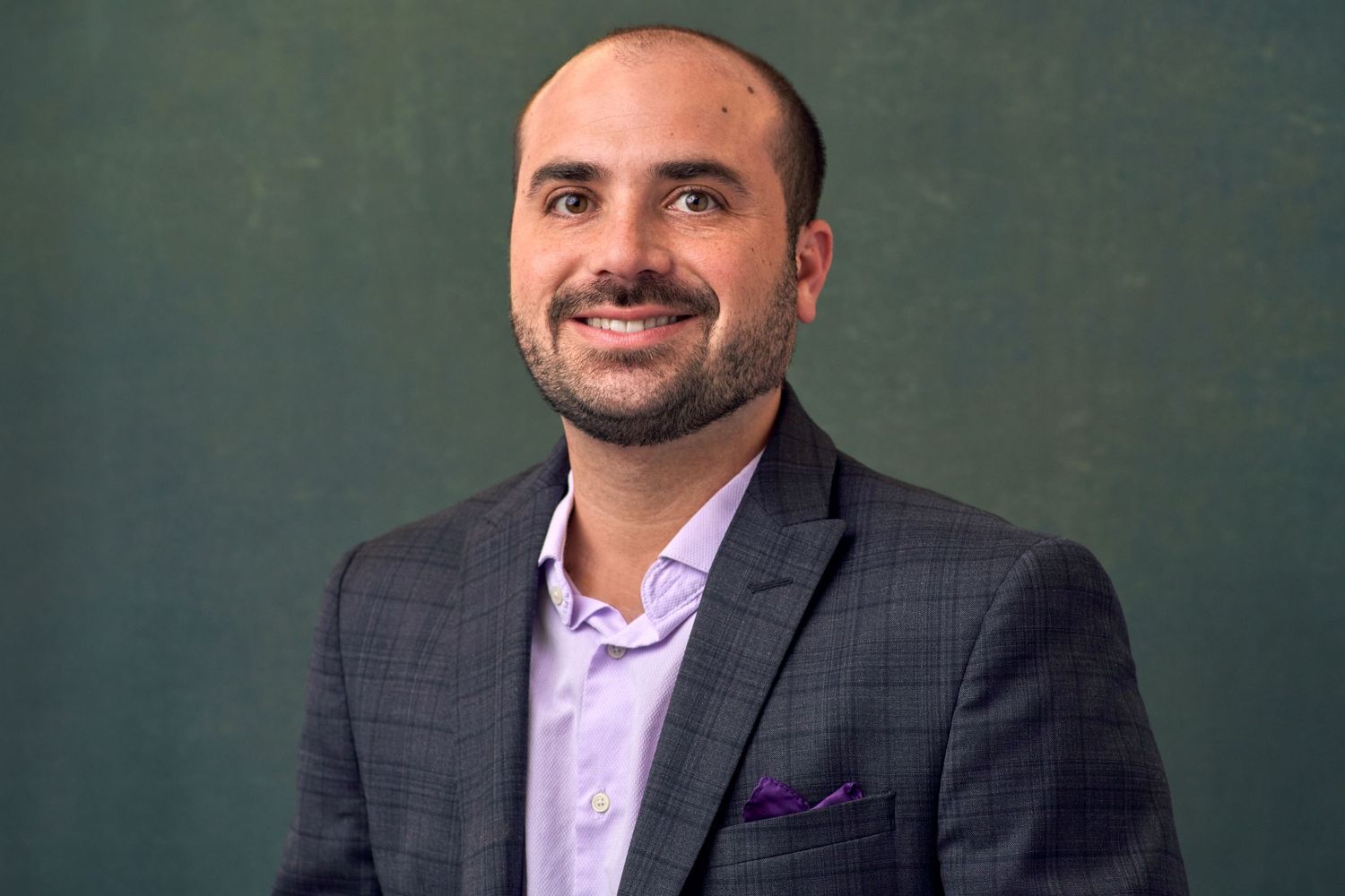 Smiling man in gray blazer and purple pocket square poses against dark green background.