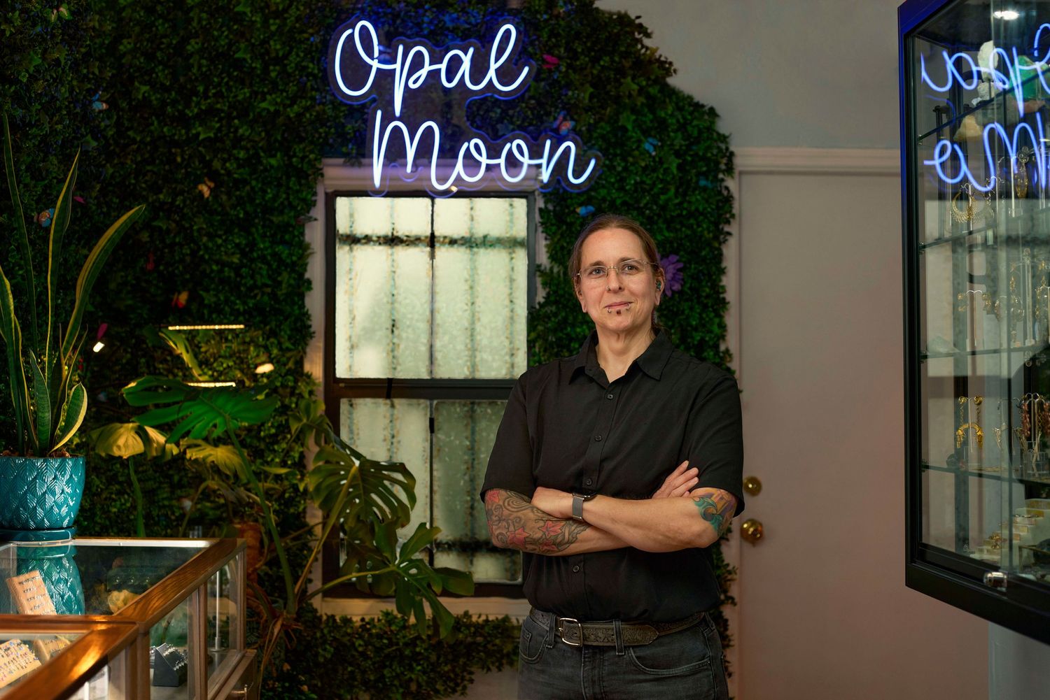 Woman with crossed arms stands before Opal Moon neon sign surrounded by lush green plants in a boutique jewelry store.