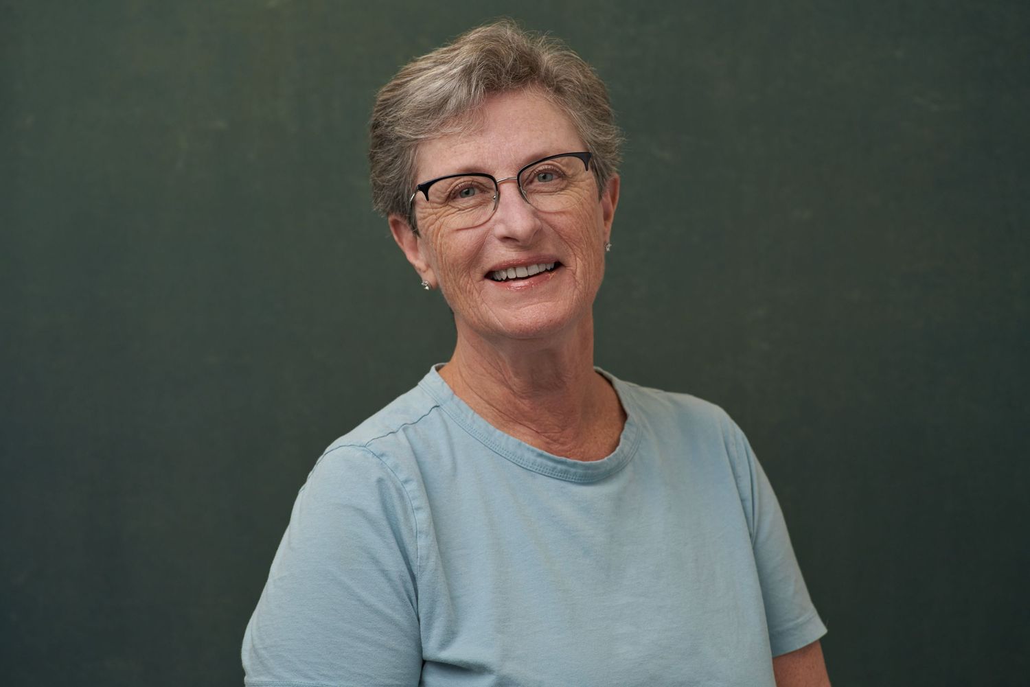 Smiling older woman with short grey hair wearing light blue shirt and glasses against green background.