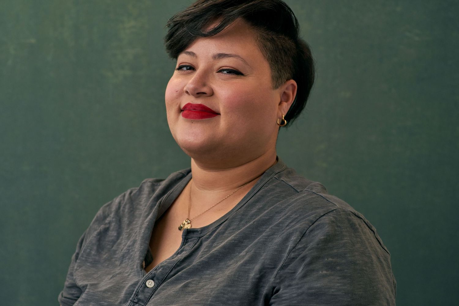 Smiling woman with short dark hair and red lips wearing grey top against dark green background.