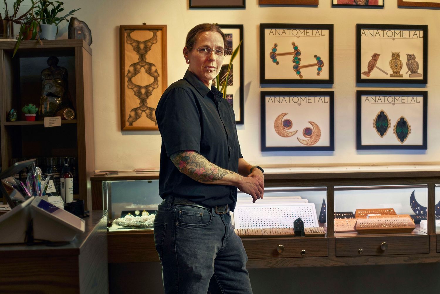 Tattooed person stands confidently in a jewelry store with display cases and framed artwork on the wall behind them.