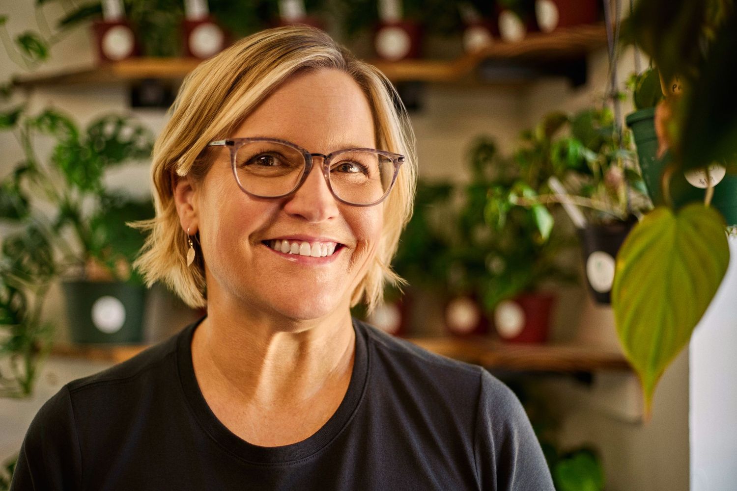 Smiling woman with glasses in a plant-filled shop wearing a black t-shirt.