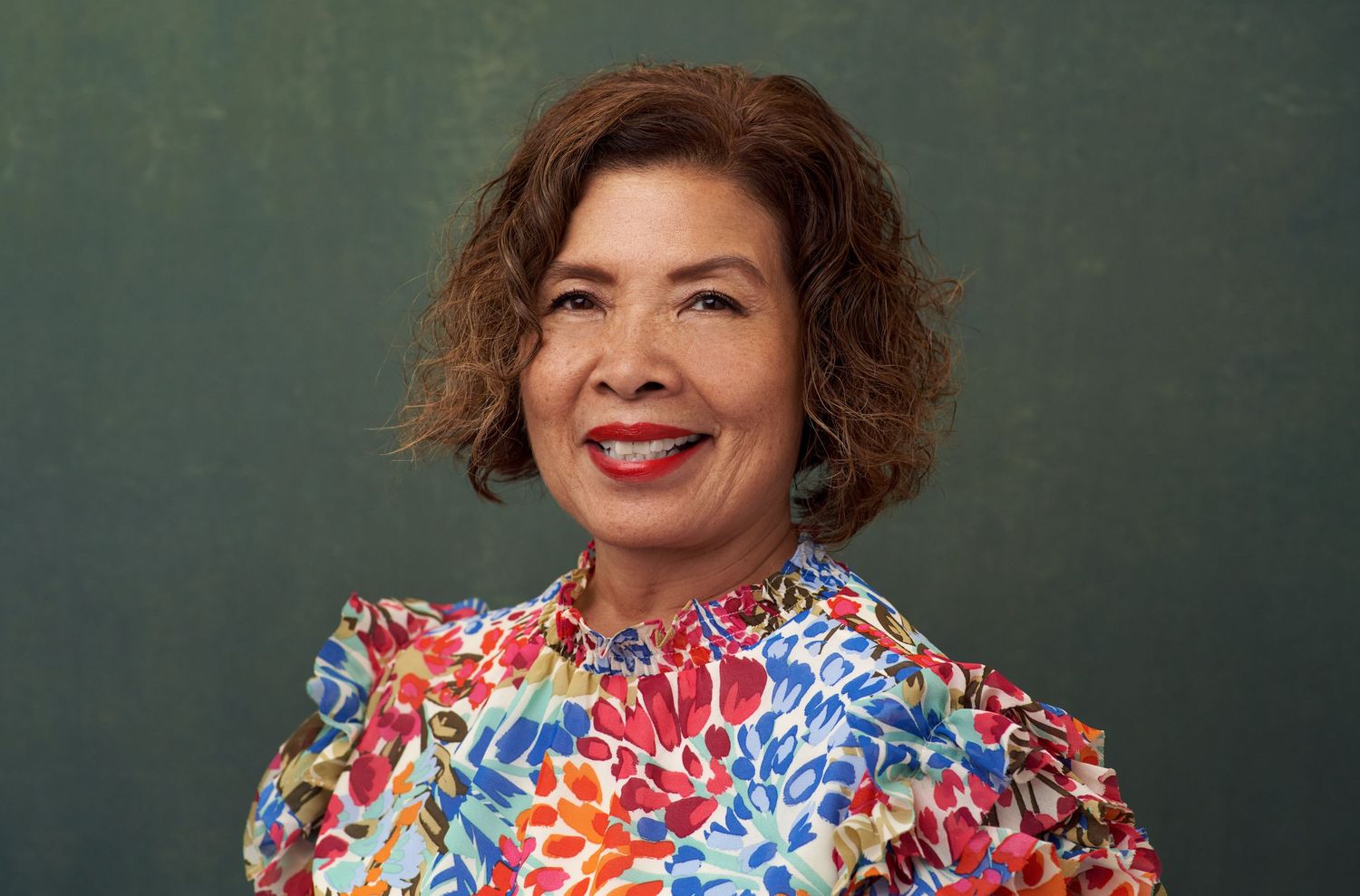 Smiling woman with curly brown hair wearing colorful floral blouse against dark green background.