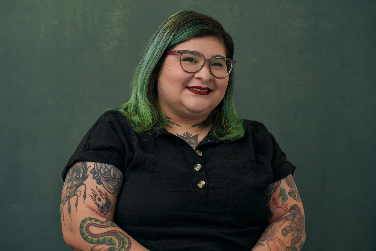 Smiling woman with green-tipped hair and tattoos wearing black top with glasses against green background.