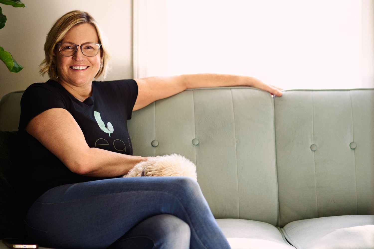 Woman in black Leaf t-shirt smiling on a mint green sofa holding a cat.