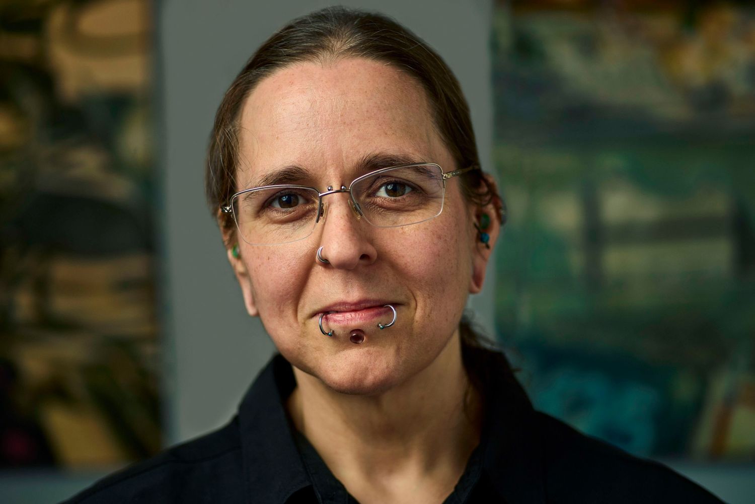 Woman with glasses and chin piercing poses in black shirt against colorful painted background.